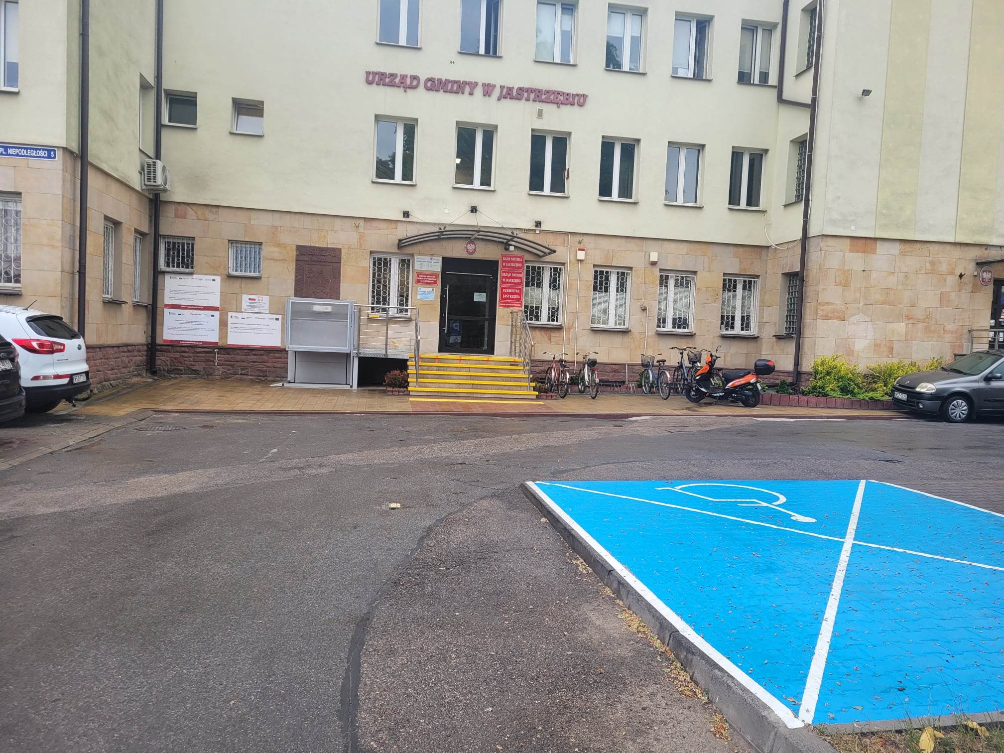 Widok wejścia do budynku Urzędu Miejskiego w Jastrzębiu w perspektywie od miejsca parkingowego dla osób z niepełnosprawnościamiInwalidów